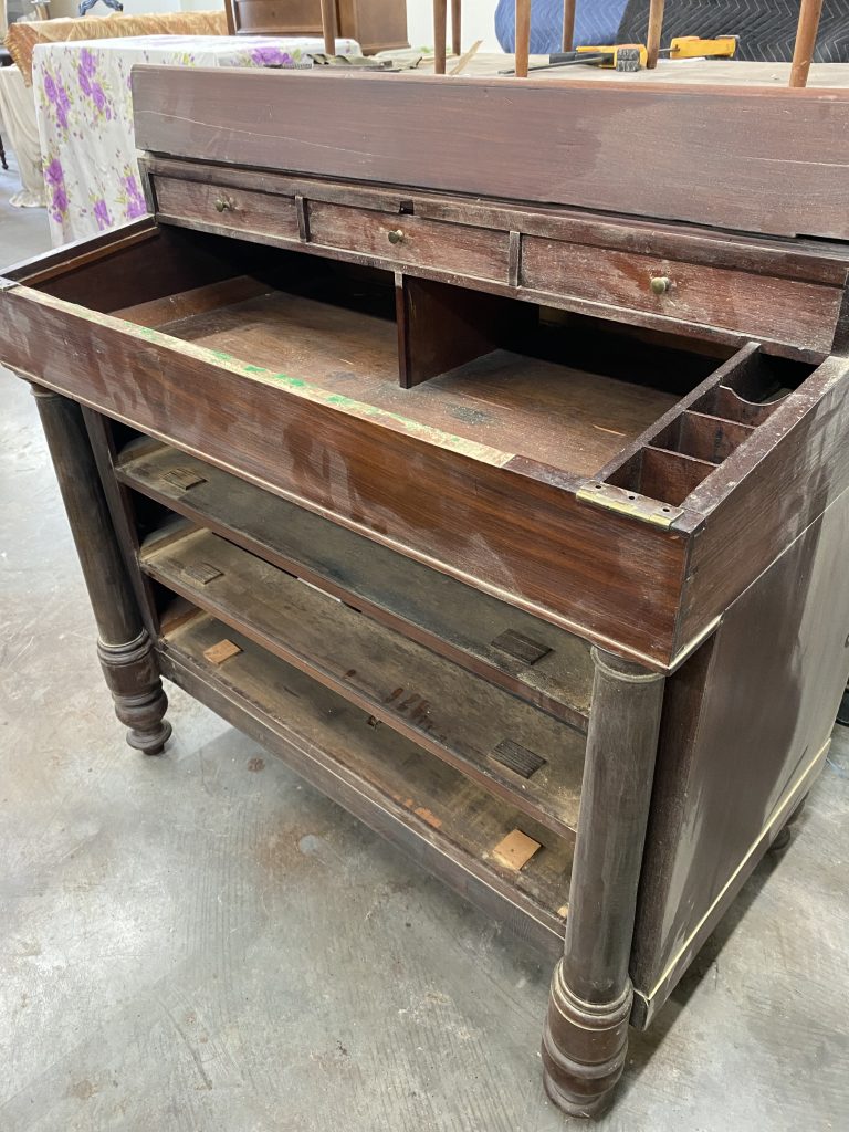 The base section of a mahogany bureau desk undergoing restoration, showcasing its intricate columns and exposed compartments as part of the repair and refinishing process.