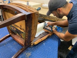 A skilled craftsperson reinforcing the underside of a chair, using a stapler to attach new padding over the wooden frame, highlighting meticulous restoration work.