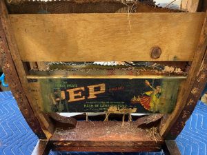 Close-up of the underside of an antique chair, revealing a repurposed vintage fruit crate panel marked “PEP” used in earlier modifications.