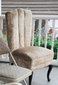 A weathered 1920s-era chair with a channeled backrest and faded upholstery, placed on a porch, showcasing its vintage aesthetic.